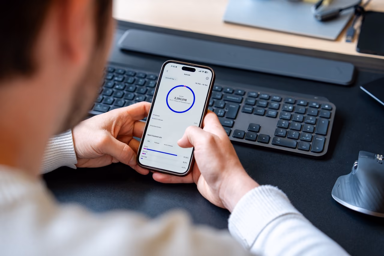 Person hält ein Smartphone in den Händen, auf dem eine App mit einem blauen Kreisdiagramm, Zahlen und Prozentangaben angezeigt wird; im Hintergrund eine schwarze Tastatur und Maus auf einem schwarzen Tisch.