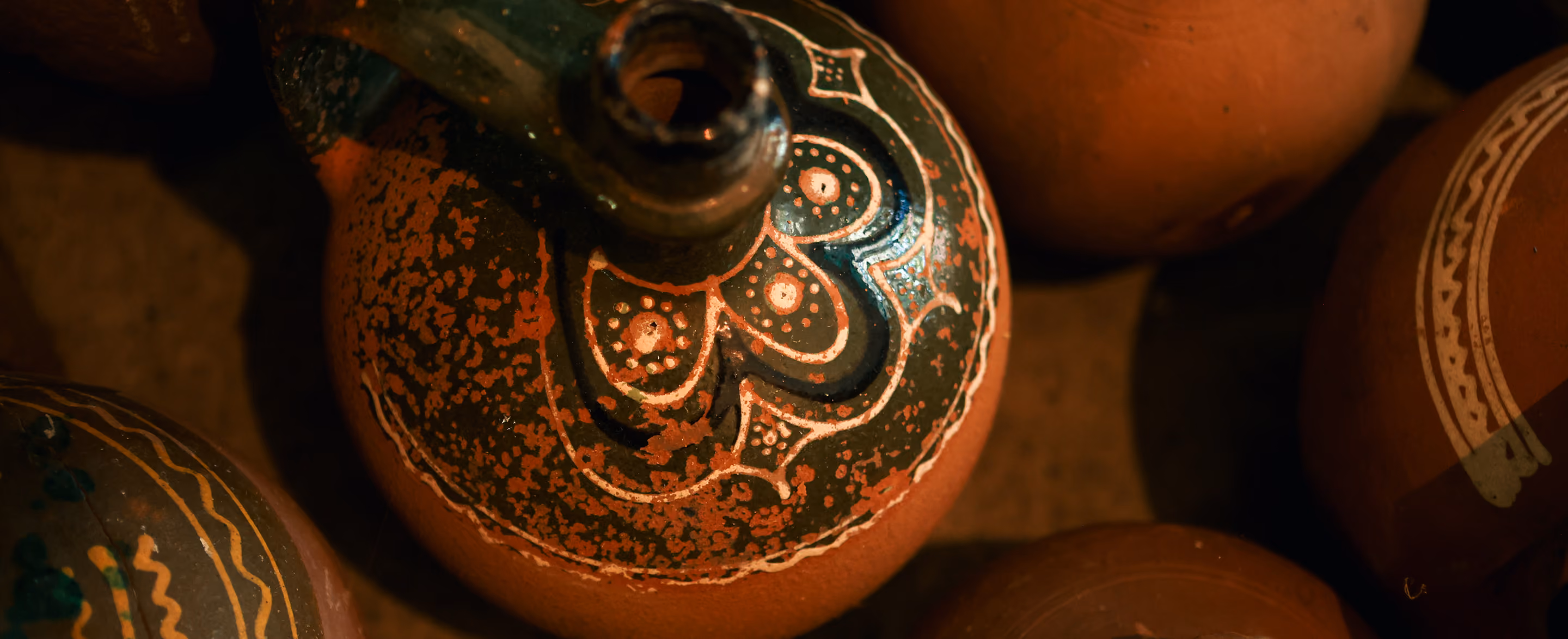 Close-up of a decorated clay pot with dark green and white painted patterns, surrounded by other plain and decorated clay pots.