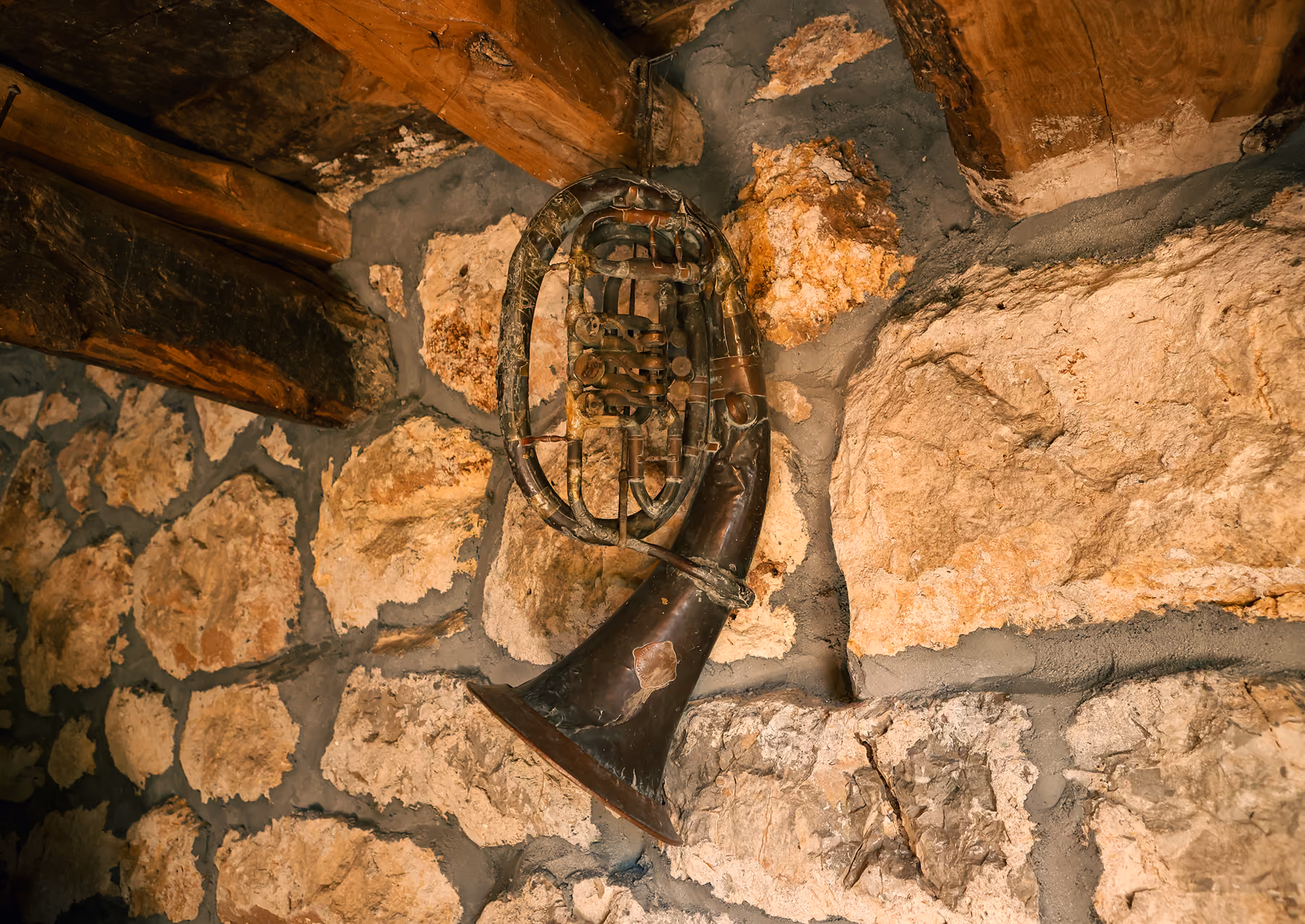 Old, tarnished brass tuba with visible dents and wear hanging on a textured stone wall with wooden beams above.
