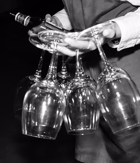 Black and white photo of a person holding four empty wine glasses upside down by their stems in one hand and a wine bottle in the other, wearing a long-sleeve shirt and vest.