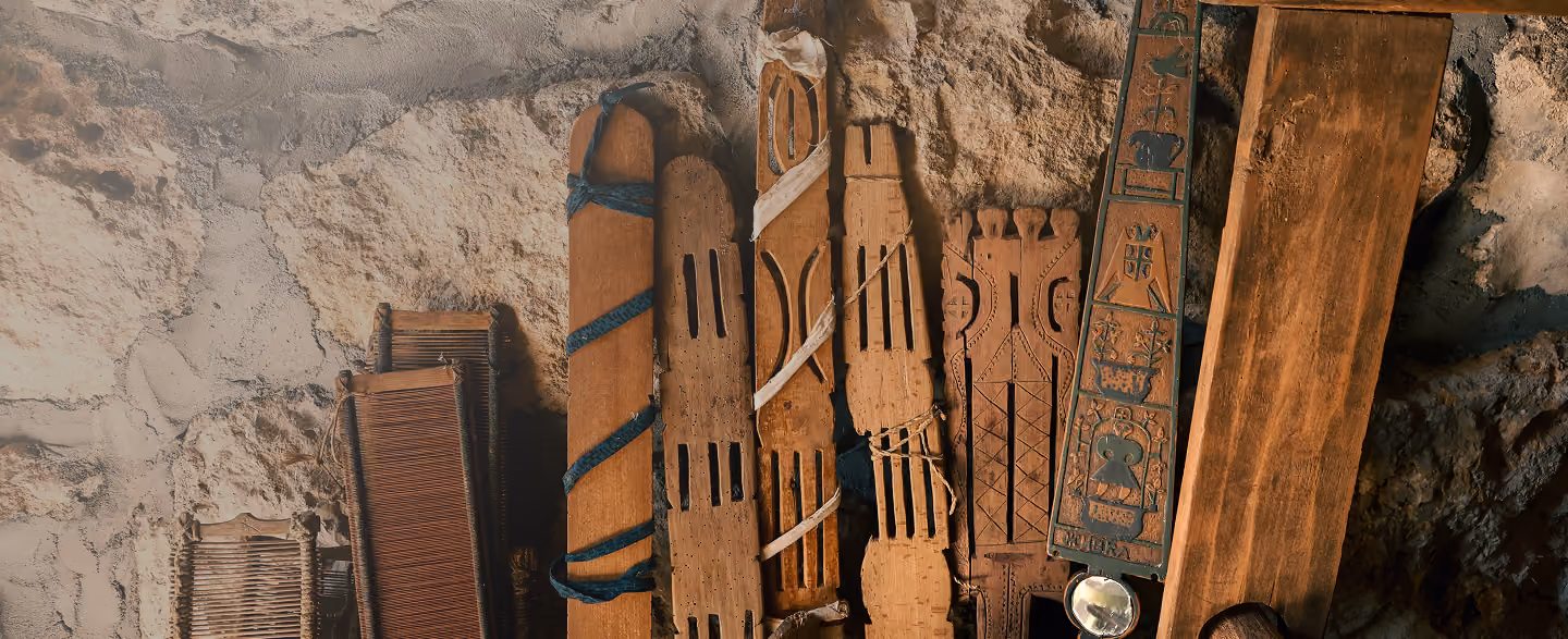 Row of various traditional wooden and woven tools or artifacts leaning against a rough stone wall, some wrapped with fabric or cord, showing different shapes and carvings.