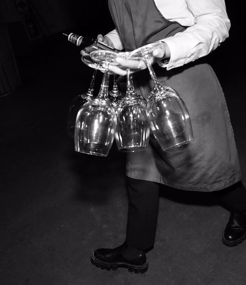 Black and white photo of a person in an apron carrying a cluster of wine glasses by their stems and holding a bottle in the other hand, walking on a dark floor.