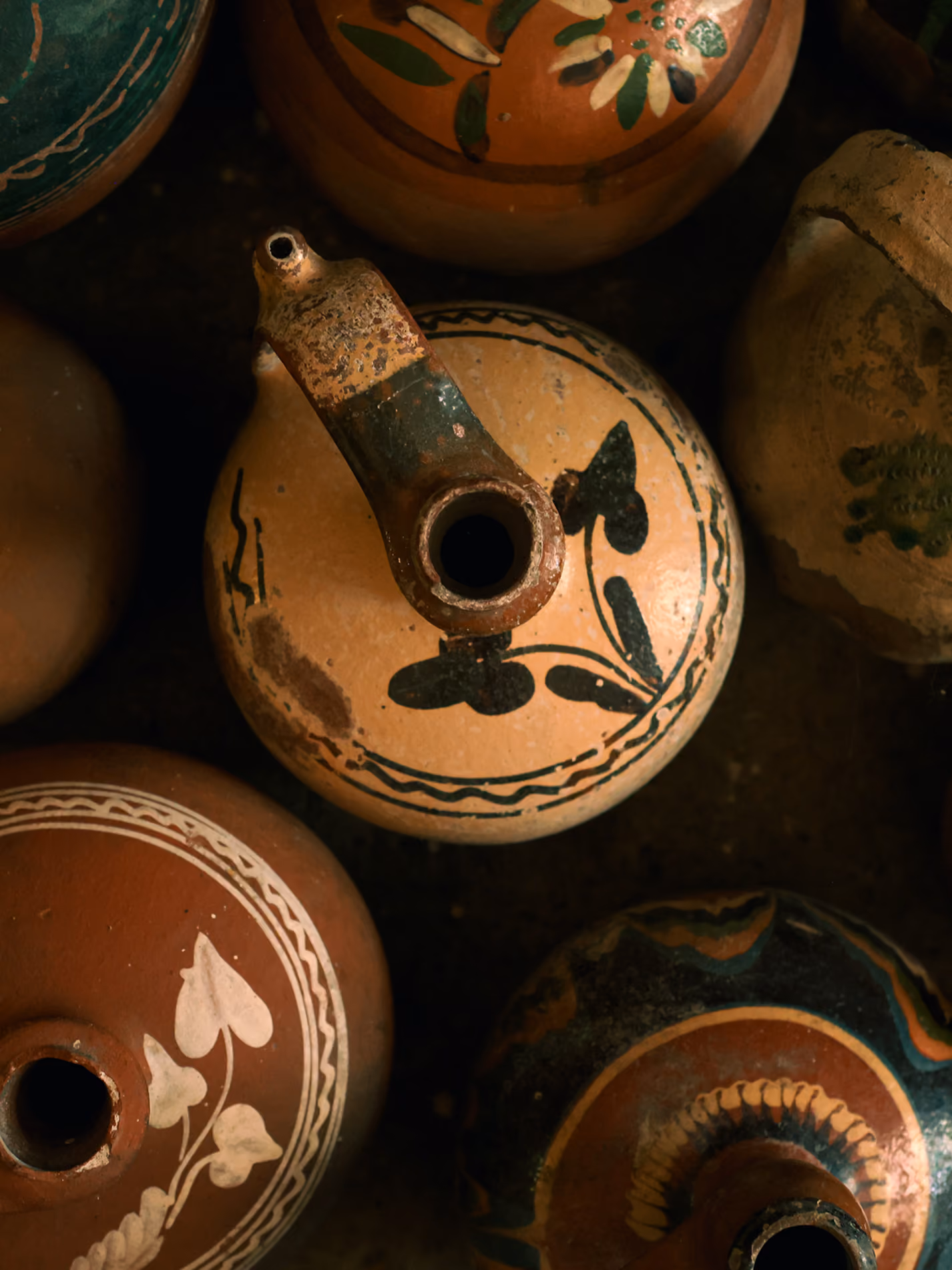 Top view of several antique ceramic vessels with spouts and painted floral and geometric designs in earthy tones.