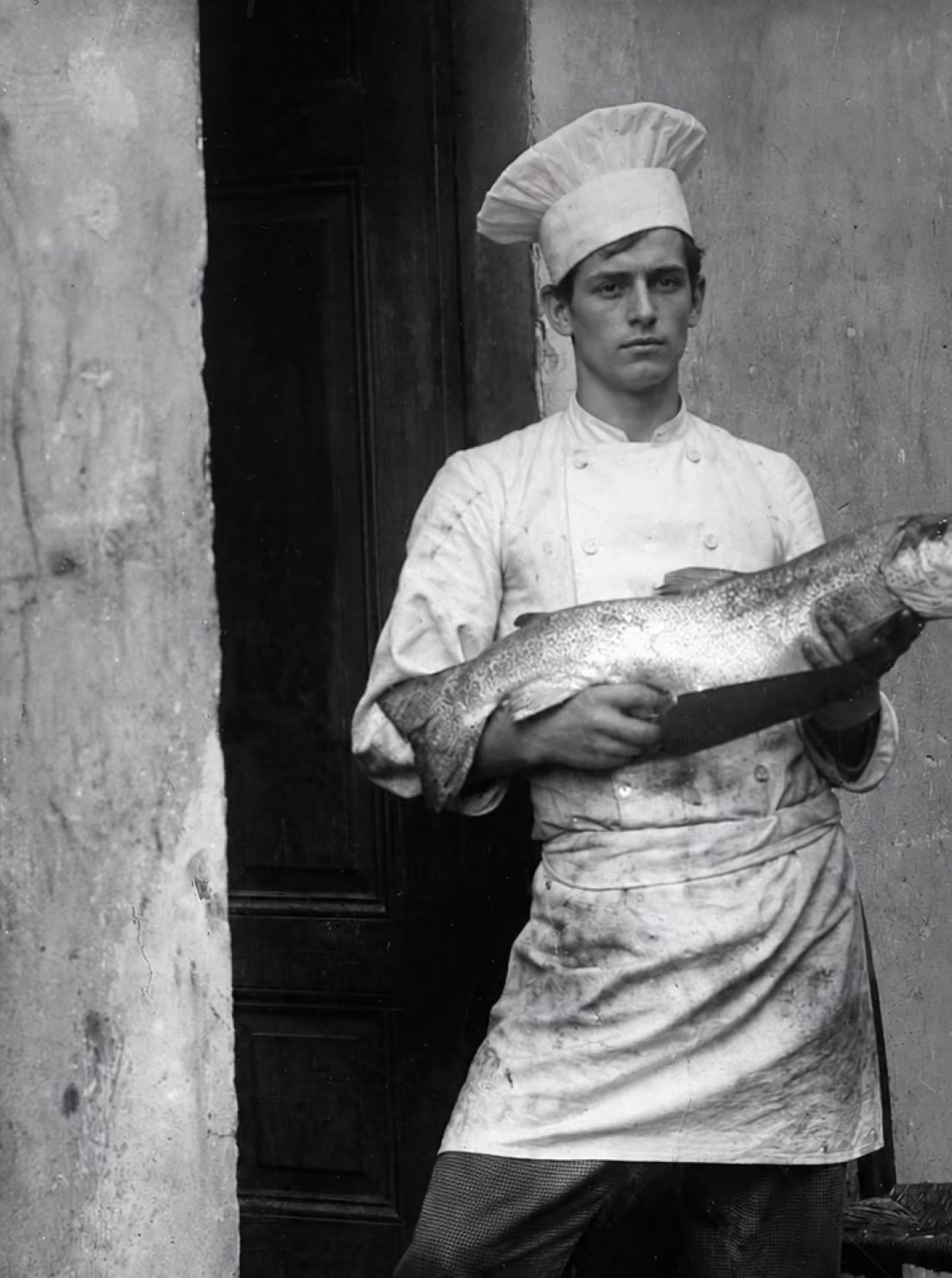 Black and white photo of a young chef wearing a white double-breasted jacket, apron, and tall chef hat, holding a large fish horizontally across his body, standing in front of a dark door and a textured light wall.