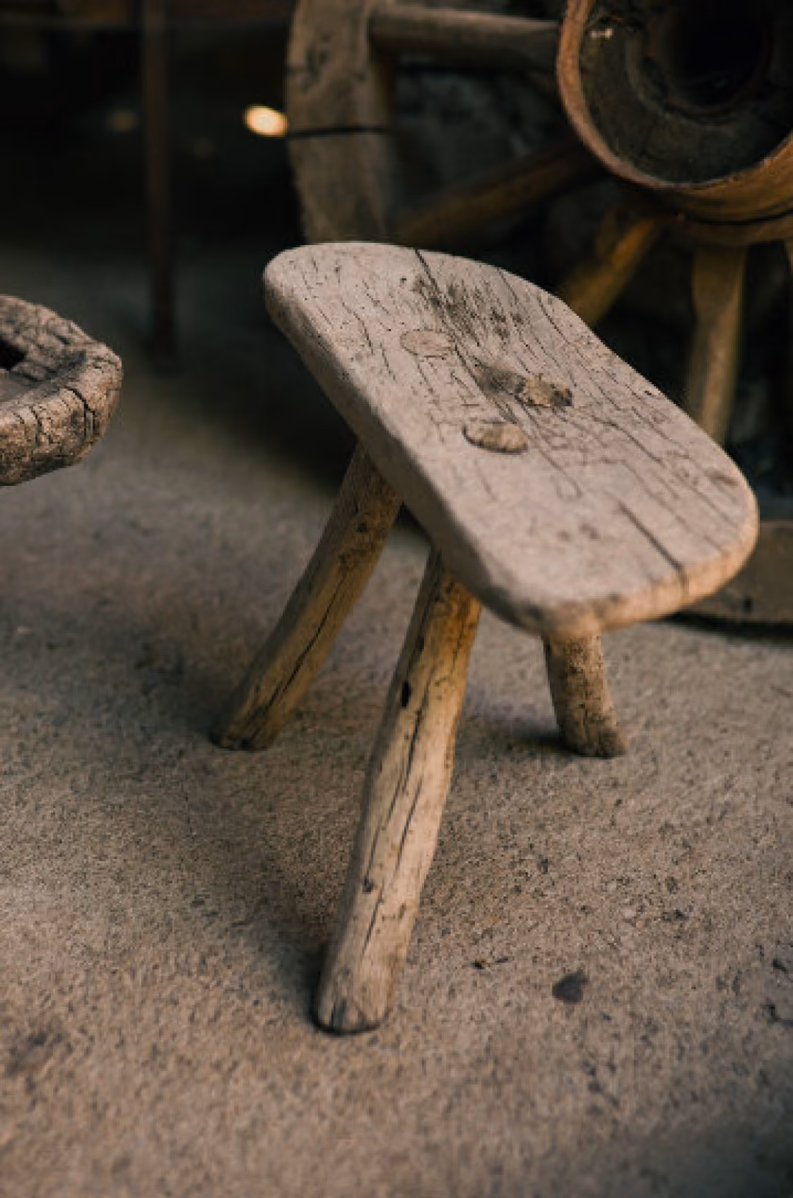 Small rustic three-legged wooden stool with a worn, cracked seat on a rough concrete floor, with old wooden objects blurred in the background.