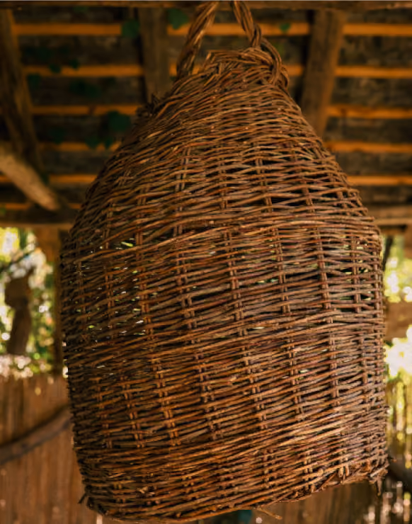 Large woven wicker basket hanging from a ceiling with wooden beams and green foliage in the background.