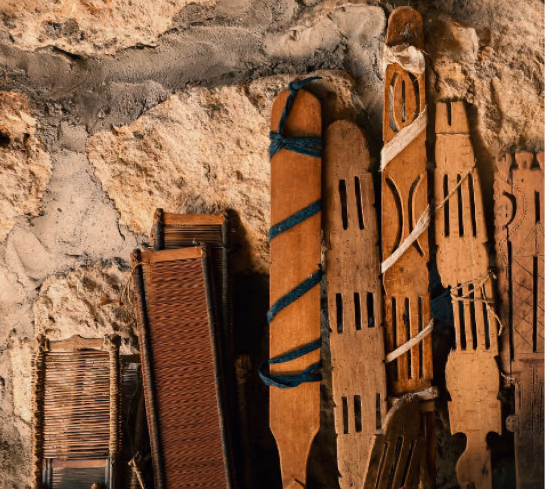 A collection of rustic wooden paddles and woven items leaned against a textured stone wall, some paddles wrapped with colorful cloth strips.