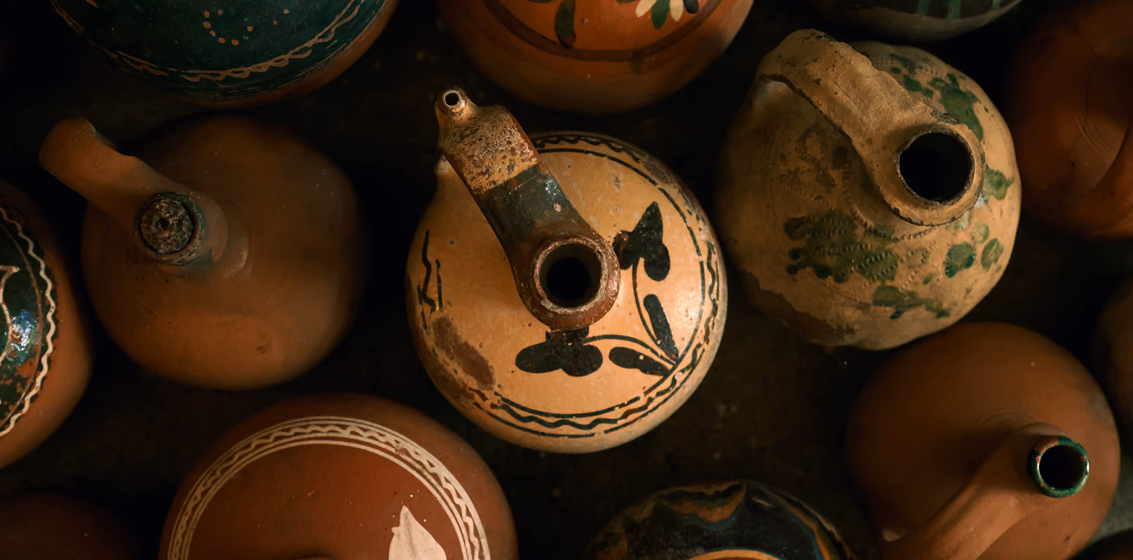 Overhead view of various traditional clay pots and pitchers with spouts, some decorated with black and green painted designs on earthy brown and beige surfaces.