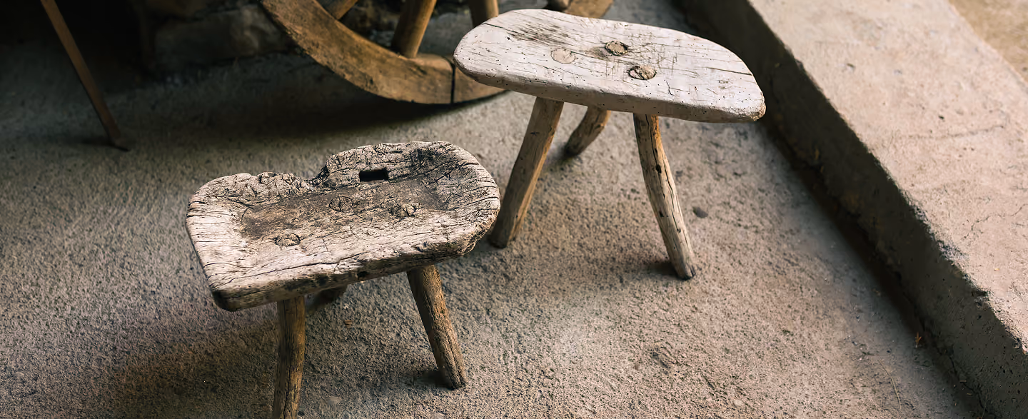 Two small rustic wooden stools with weathered surfaces and three legs each, placed on a rough concrete floor near stone steps.