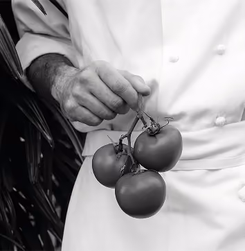 Chef in white uniform holding a cluster of three tomatoes by their stems.