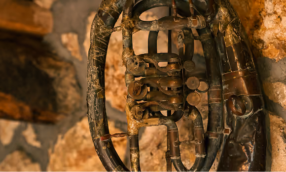 Close-up of an old, worn brass musical instrument with valves and circular tubing, set against a rustic stone wall background.