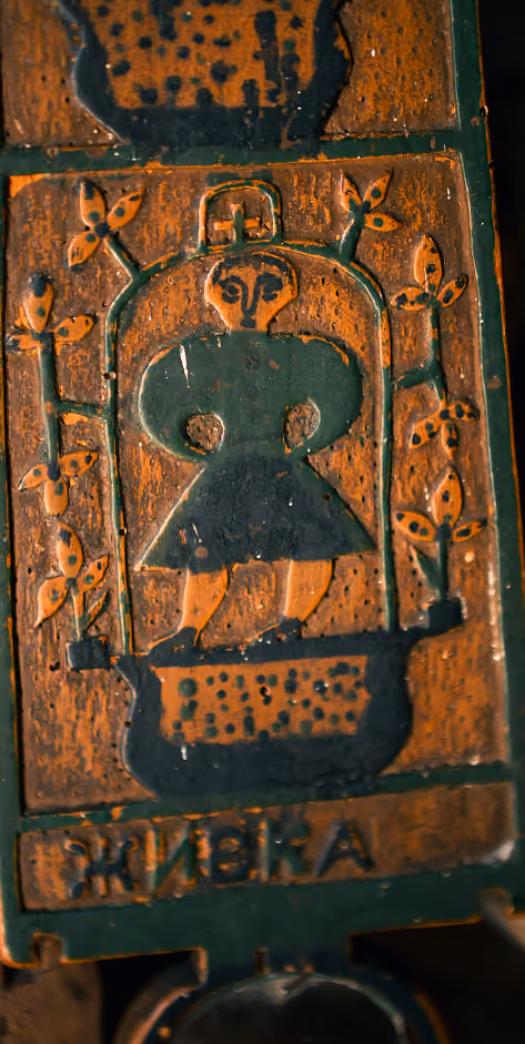 Rusty metal plate with an embossed figure of a person wearing a dress, surrounded by floral designs, with Cyrillic text at the bottom.