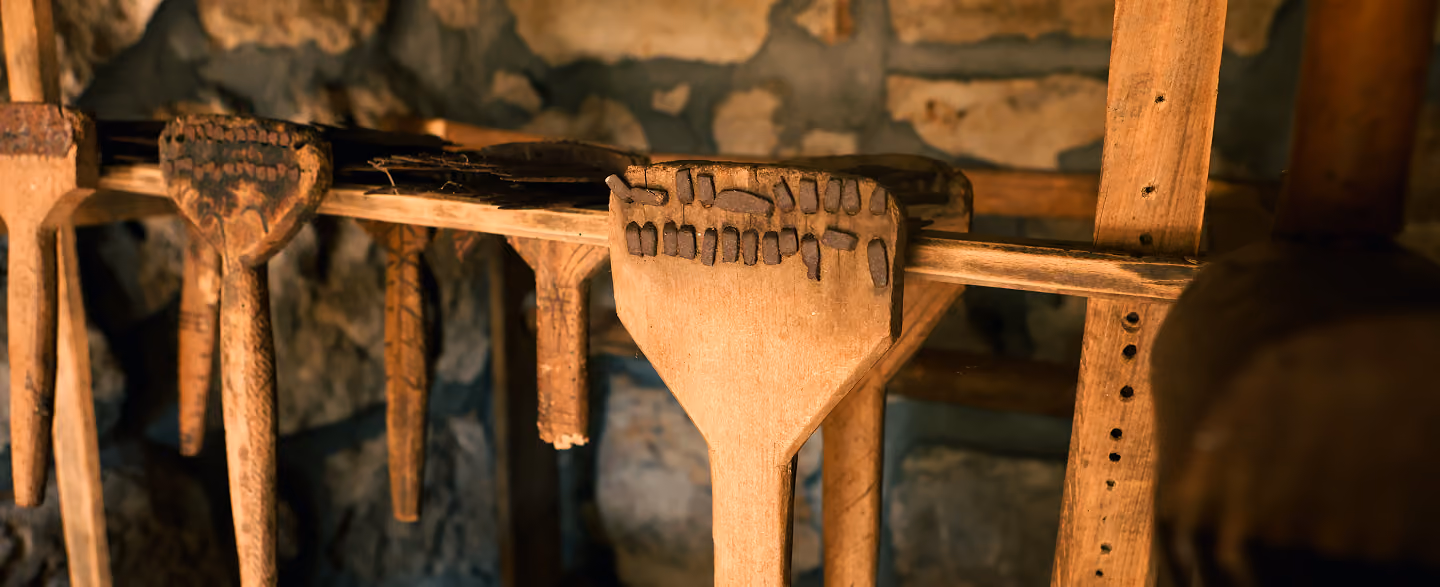Close-up of traditional wooden combs and tools hanging on a wooden rack against a stone wall background, softly lit by warm light.