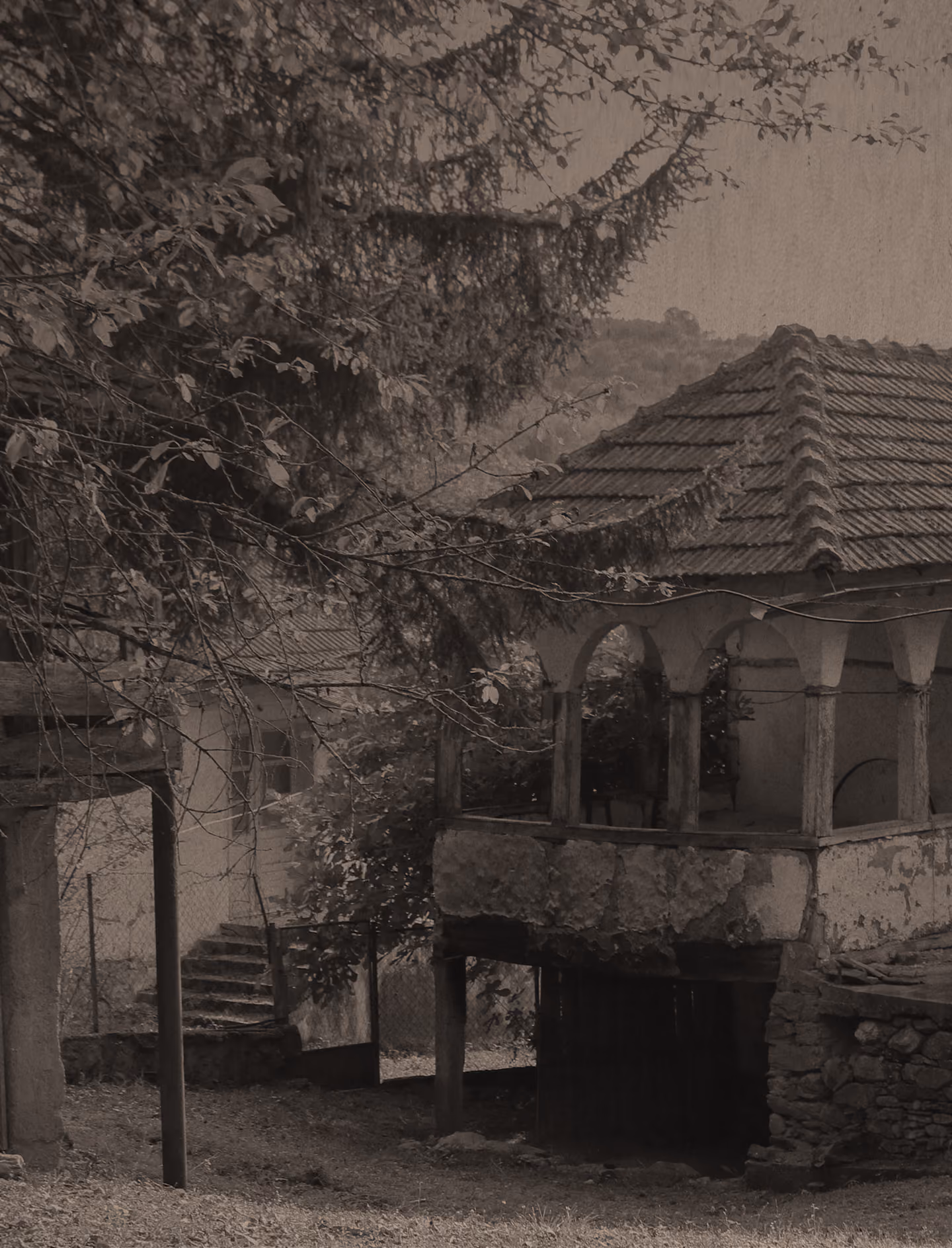 Sepia-toned image of an old stone building with a tiled roof and an open balcony supported by four columns, surrounded by trees and foliage, with stone steps visible in the background.