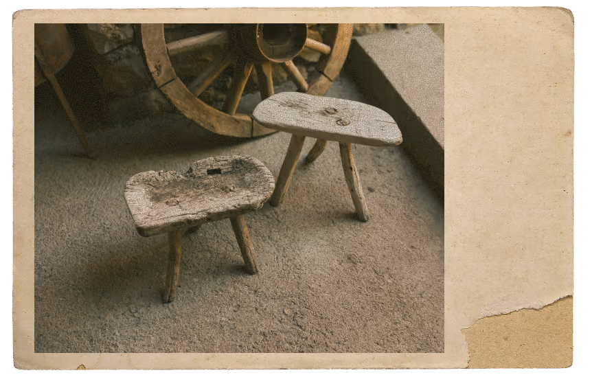 Two small rustic wooden stools with three legs each, placed on a rough surface near a stone wall with a large wooden wagon wheel.