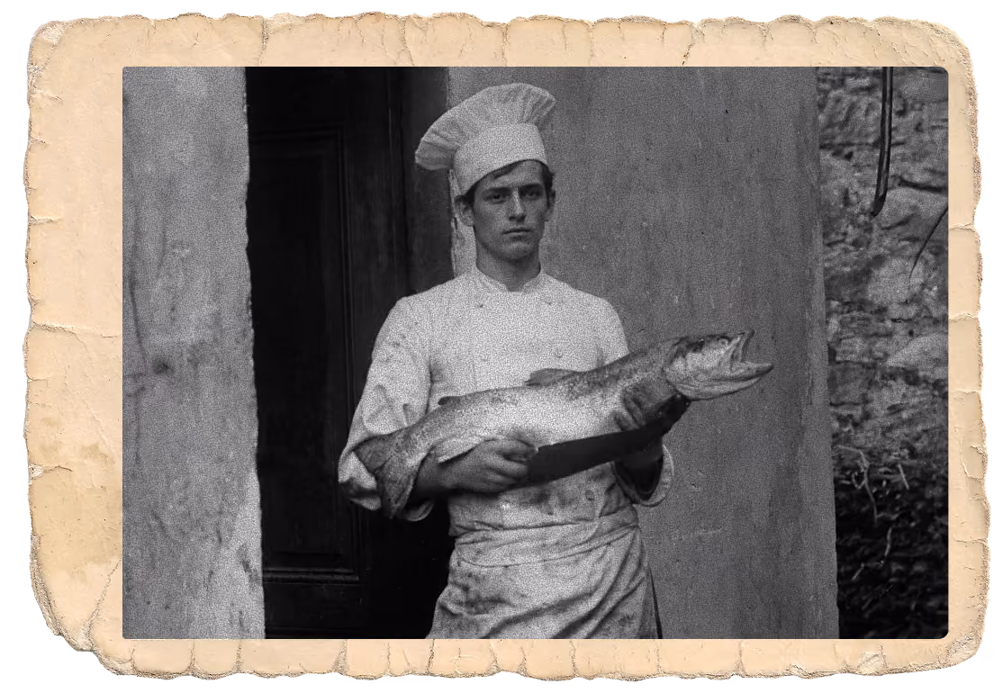 Black and white vintage photograph of a young chef wearing a tall chef hat and double-breasted coat, holding a large fish in both hands, standing inside a doorway with stone walls.