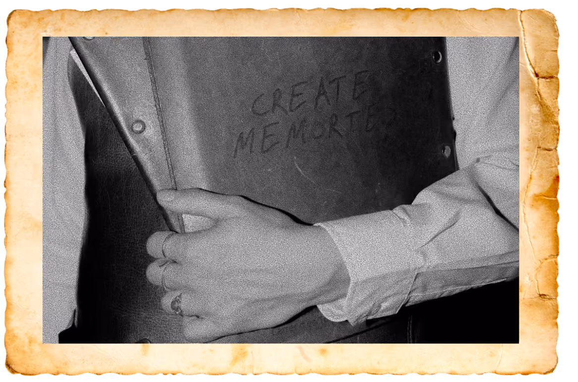 Black and white photo of a person wearing a buttoned shirt holding a dark book or folder with the handwritten words 'CREATE MEMORIES' on the cover. The photo has a vintage, worn paper border.