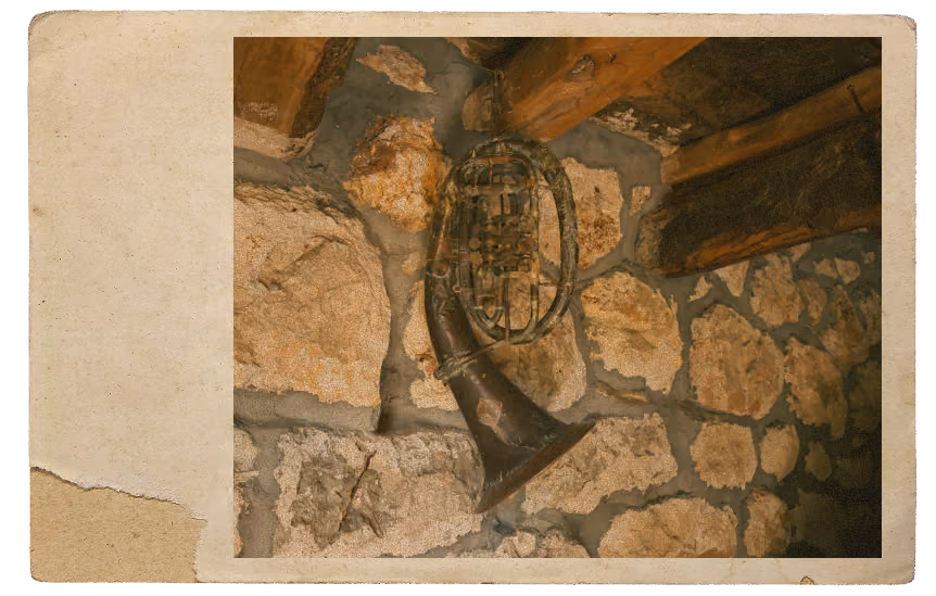 Old brass musical instrument, possibly a tuba or euphonium, hanging on a stone wall inside a rustic wooden-beamed room.