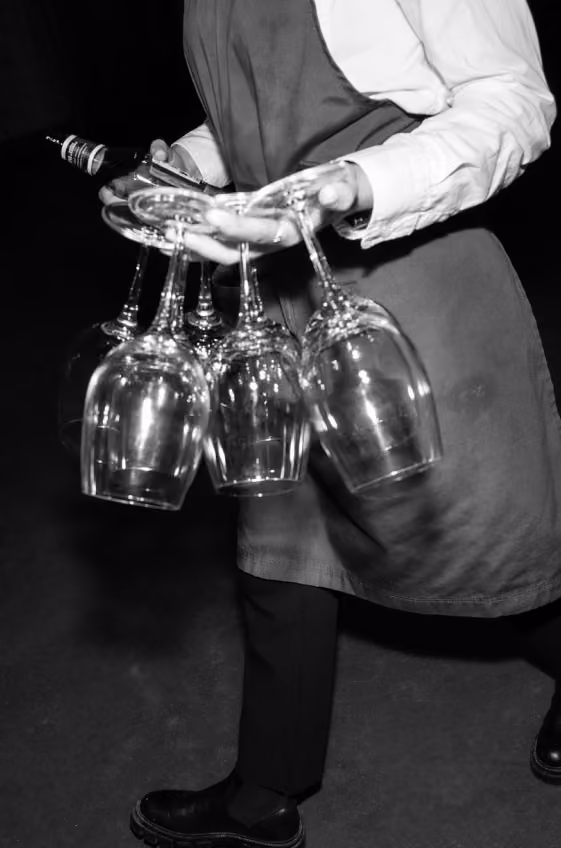 Person in apron and white shirt holding several upside-down wine glasses in one hand and a bottle in the other, walking on a dark floor.
