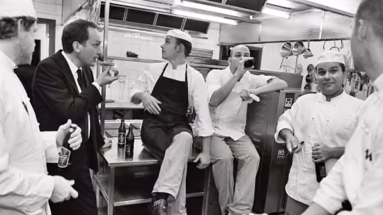 Black and white photo of six men casually socializing in a professional kitchen, some wearing chef uniforms and hats, others in regular clothing, holding bottles and sitting or standing around kitchen equipment.