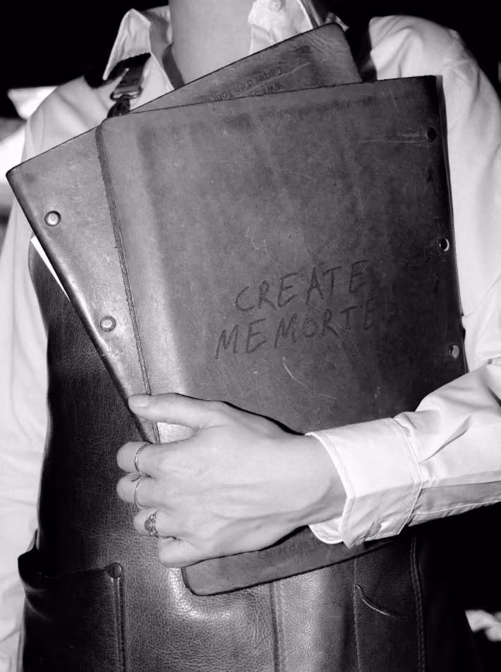 Person wearing a white shirt and dark leather apron holding two vintage binders, one with the handwritten phrase 'CREATE MEMORIES'.