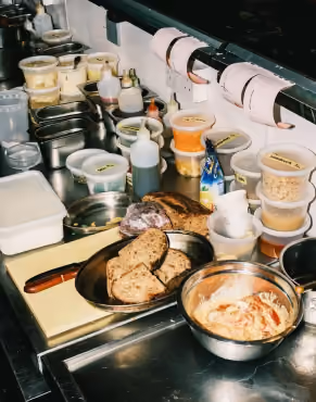 Kitchen prep station with sliced rustic bread on a tray, a bowl of mixed ingredients, condiment bottles, multiple stacked containers with ingredients, and printed order tickets hanging above the counter.