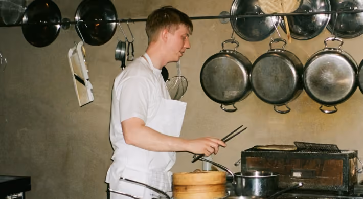 A chef in a white shirt and apron uses tongs to prepare food in a kitchen with hanging pans and cooking utensils.