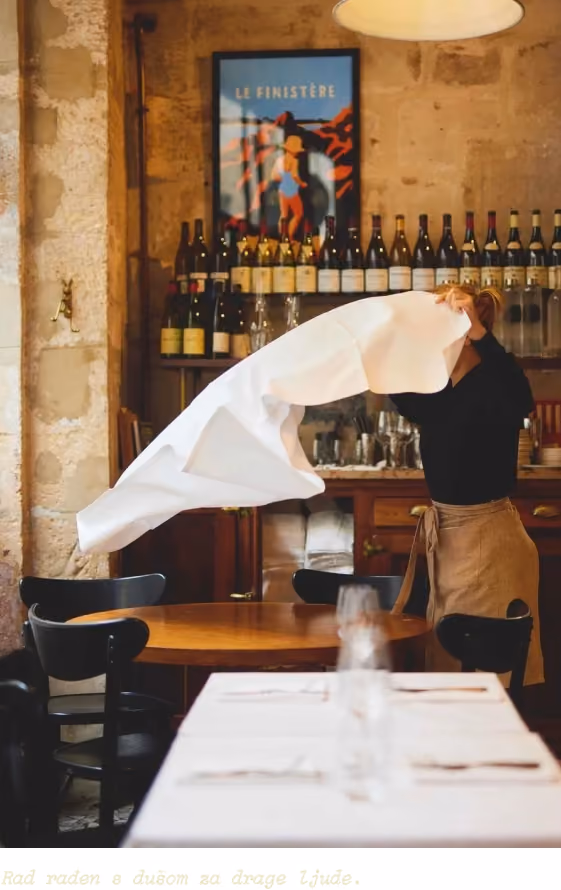 A person wearing a black top and brown apron spreads a white tablecloth over a round wooden table in a cozy restaurant with stone walls, wooden chairs, a wine bottle display, and a framed Le Finistère poster.