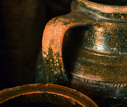 Close-up of a rustic ceramic jug with a curved handle, featuring a textured, mottled glaze in earthy tones of brown, green, and orange.