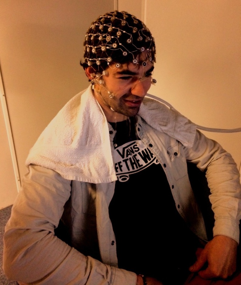 Young man wearing a beige jacket and a black Vans t-shirt sitting with an EEG cap with electrodes on his head, connected to wires, and a white towel draped over his shoulders.