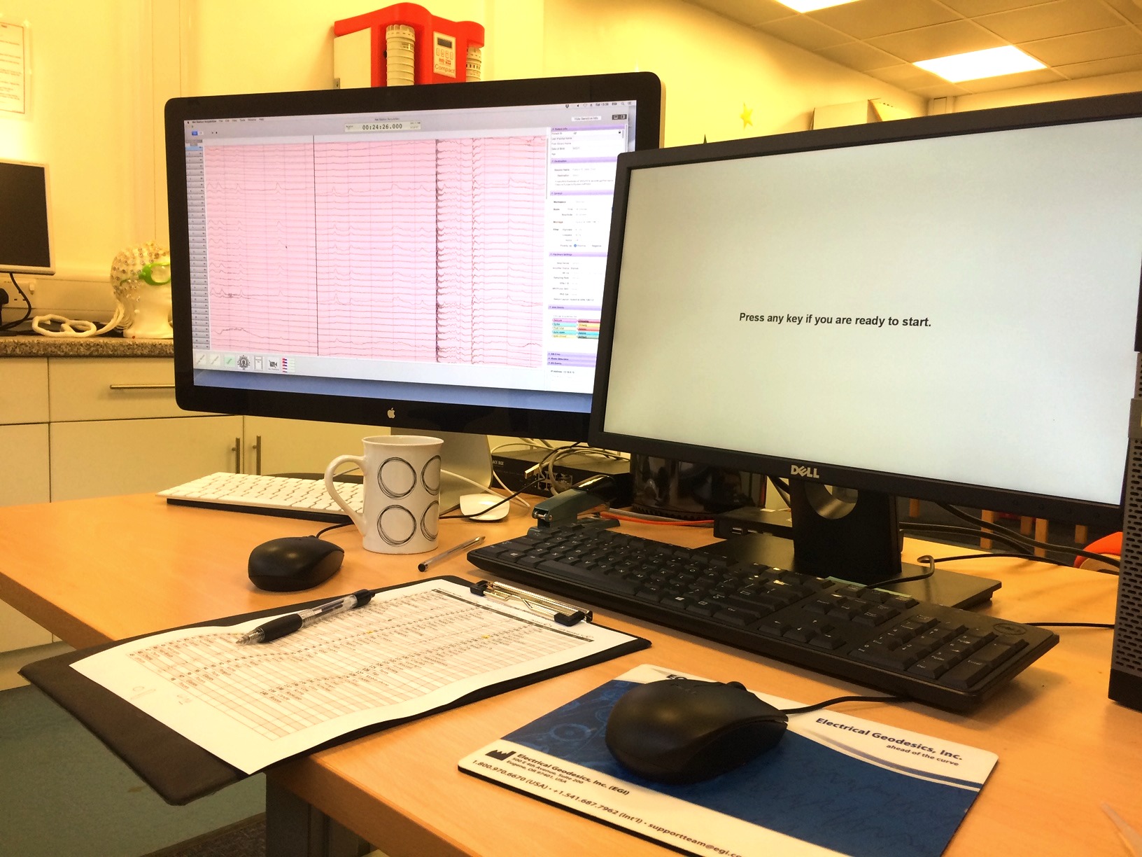 Desk with two computer monitors: the left screen shows brain wave data with multiple lines on a pink background, and the right screen displays the text 'Press any key if you are ready to start.' A keyboard, two computer mice, a clipboard with a printed data sheet and pen, and a white mug with circle designs are on the desk. In the background, there is a counter with a white EEG skull cap and other lab equipment.