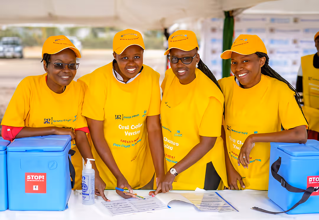 Four health workers in yellow oral cholera vaccine T-shirts and caps smiling behind vaccine cool boxes