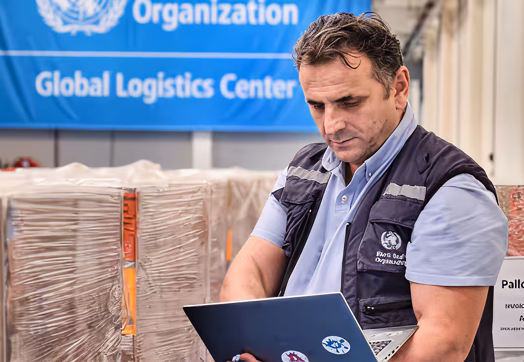 WHO worker using a laptop in front of stacked supplies at the WHO Global Logistics Center