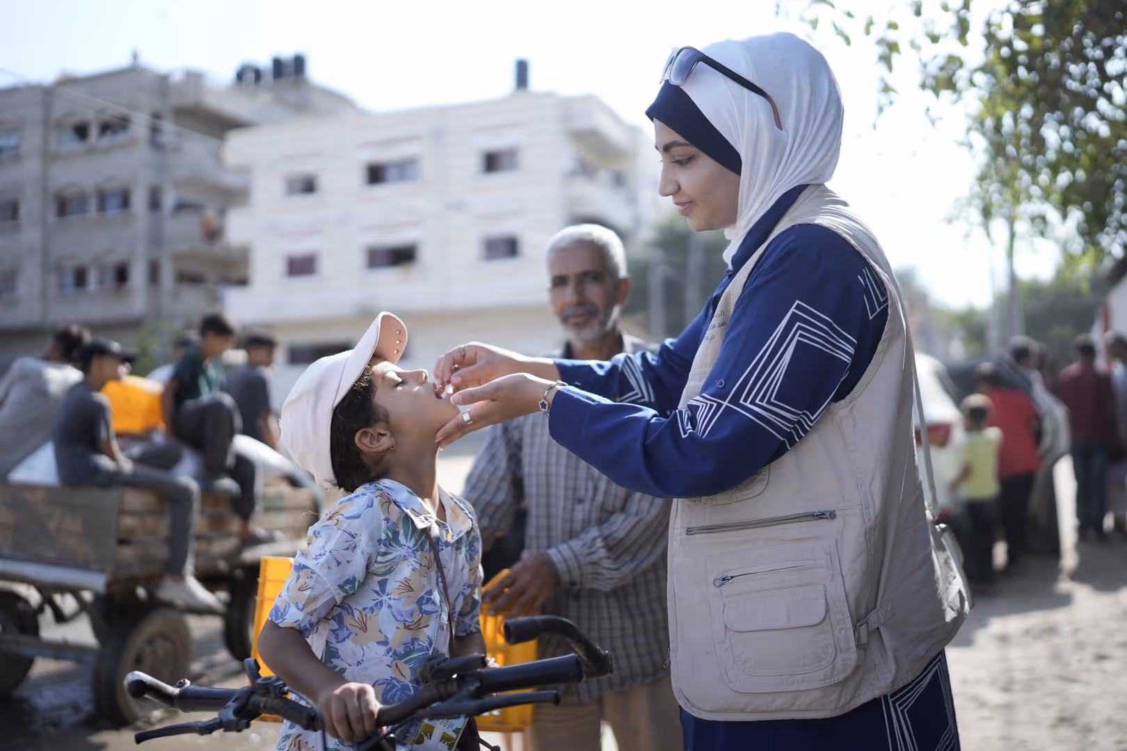 Woman in a headscarf and WHO vest giving oral vaccine drops to a child in a busy street