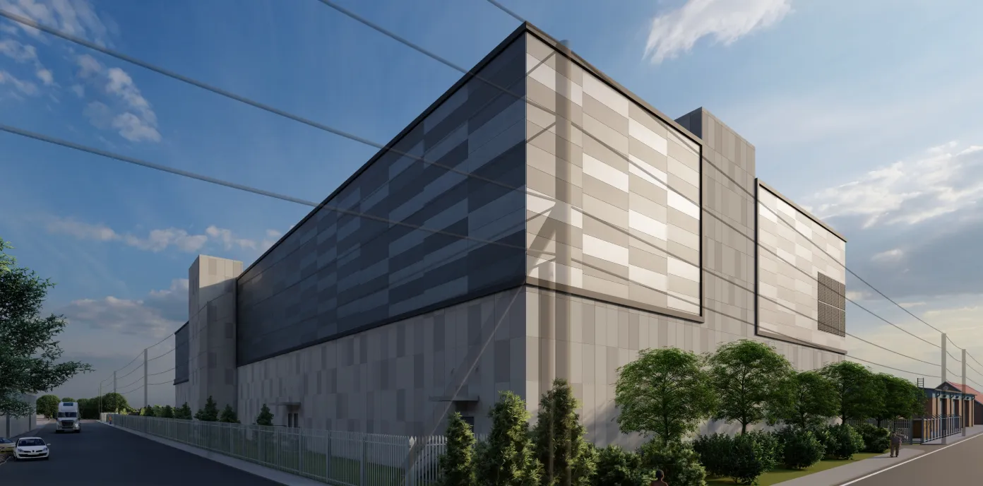 A large modern industrial or commercial building with a neutral-toned facade and large rectangular panels, surrounded by a metal fence and greenery, under a partly cloudy sky during daylight.