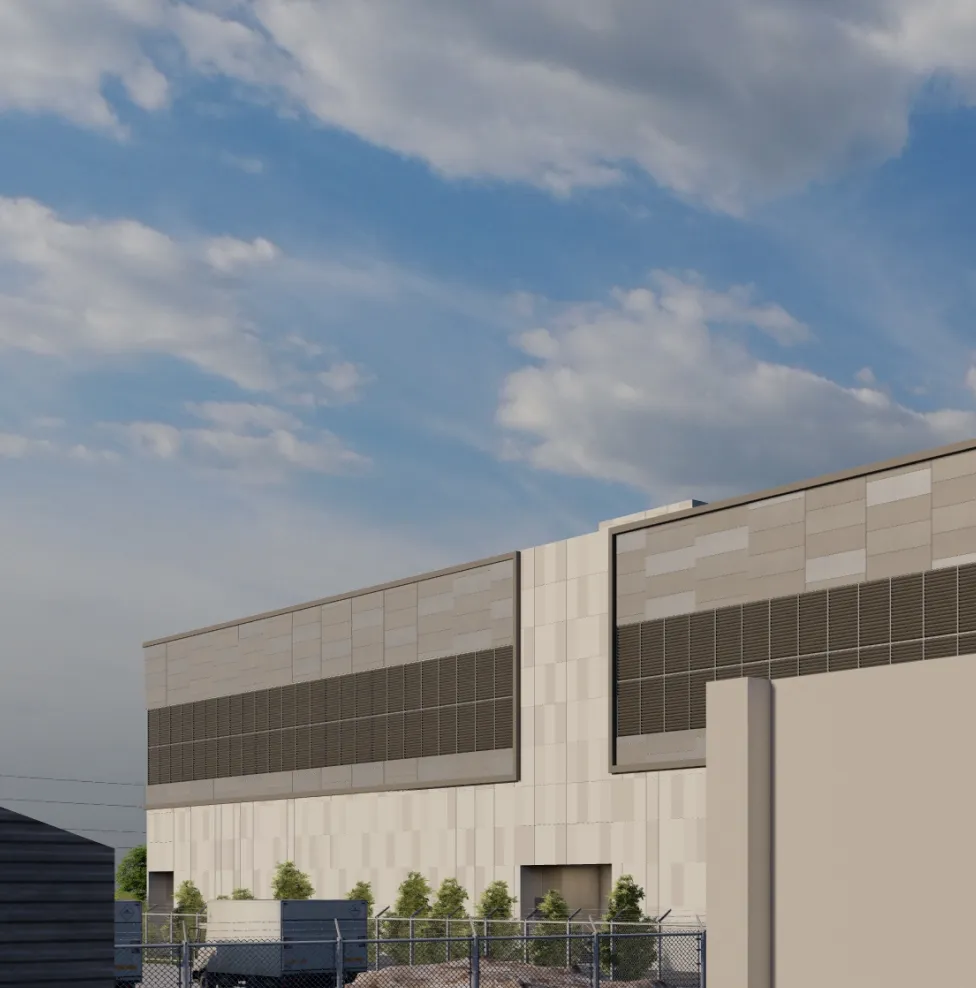 Modern industrial building with beige panel walls and large dark ventilation sections near the roof under a partly cloudy blue sky, surrounded by a chain-link fence and some vehicles.