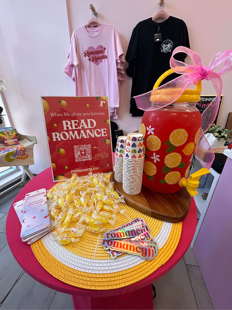 A close-up of a event table featuring a large glass jar of red lemonade wrapped with a pink ribbon, paper cups, yellow lemon-flavored candies, stickers, and a red framed sign reading "When life gives you lemons READ ROMANCE".