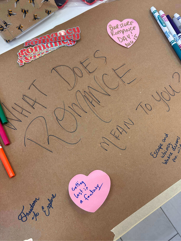 A brown paper sheet labeled "WHAT DOES ROMANCE MEAN TO YOU?" written in large marker, featuring heart-shaped sticky notes with handwritten thoughts like "Getting lost in a fantasy" and "Book Store Romance Day! Aug 15".