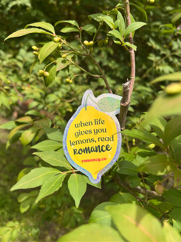 A lemon-shaped glittery sticker placed on a tree branch among green leaves, reading "when life gives you lemons, read ROMANCE" with "romancy.co" printed underneath.