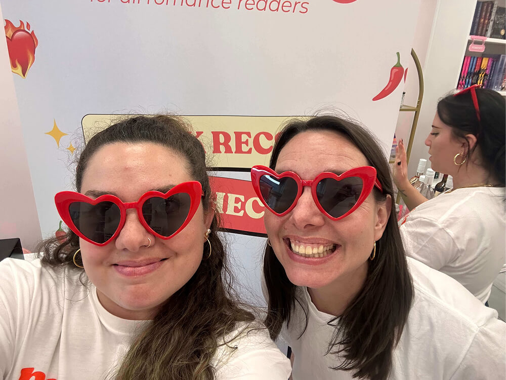 Two smiling women wearing matching red heart-shaped sunglasses and white t-shirts pose for a close-up selfie. In the background, a third person is partially visible near a display board with graphics of hot sauce peppers.