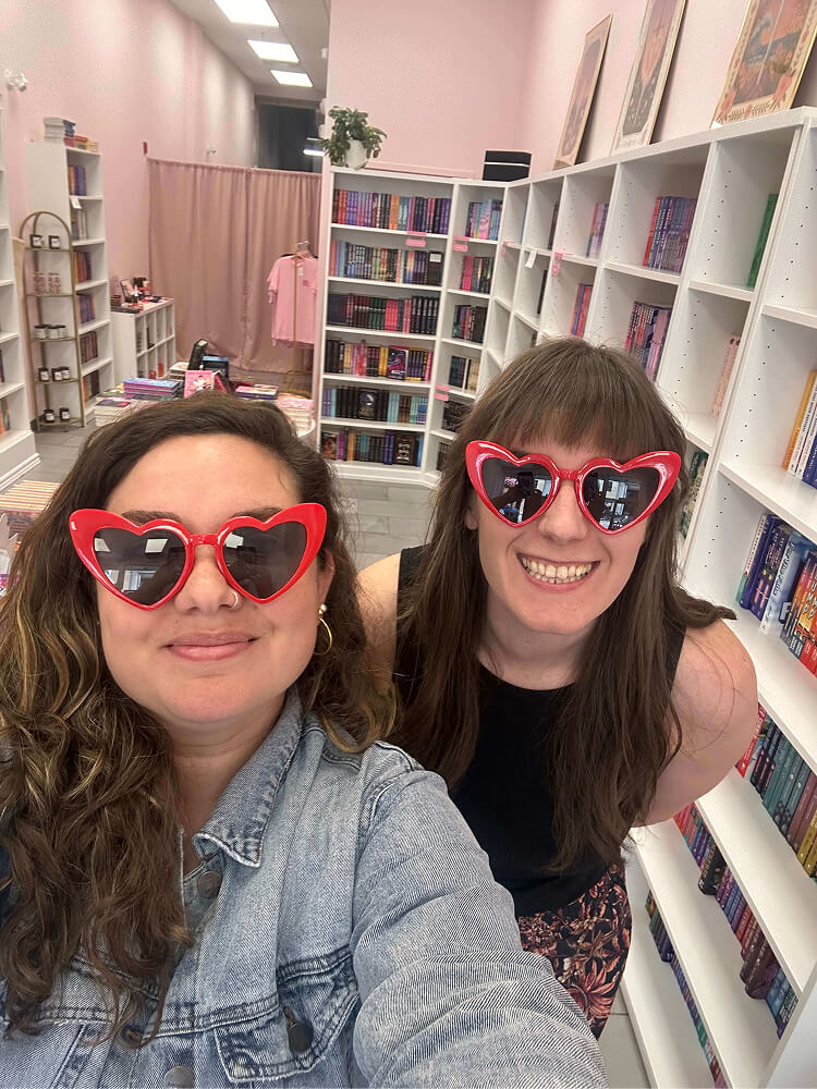 Two women wearing matching red heart-shaped sunglasses smile for a selfie inside a bookstore filled with white bookshelves packed with books.