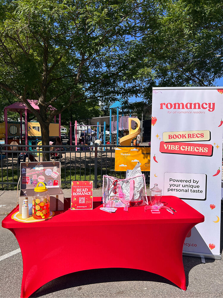 A bright red table setup outdoors in front of a playground, featuring a large banner for "romancy: FOR ALL ROMANCE READERS - BOOK RECS & VIBE CHECKS". The table holds a lemonade dispenser, candy display trunk, promotional sign, and a clear tote bag with heart sunglasses.