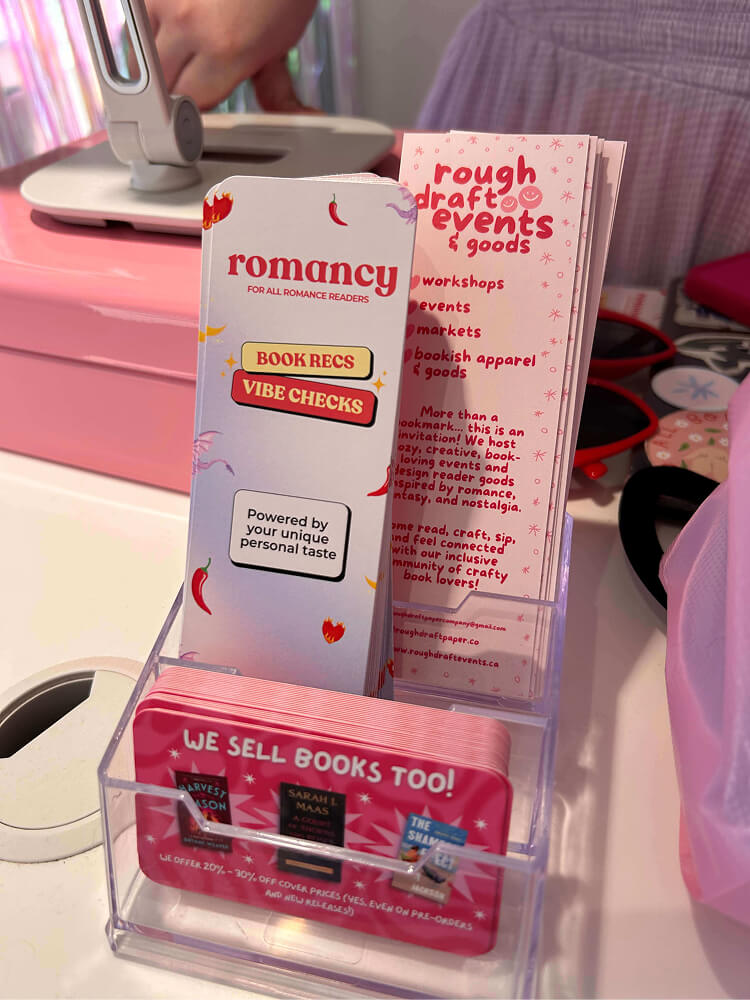 clear acrylic desk organizer holding bookmarks and event flyers. The front card reads "WE SELL BOOKS TOO!", and a bookmark displays "romancy: FOR ALL ROMANCE READERS" with buttons for "BOOK RECS" and "VIBE CHECKS".