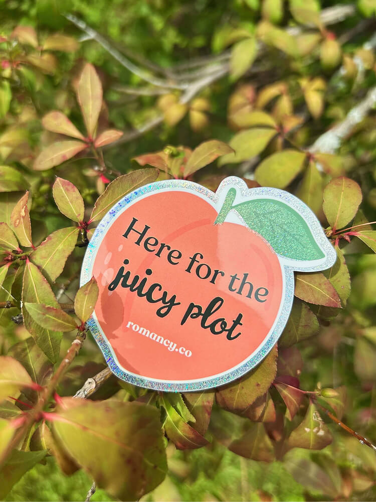 A peach-shaped glittery sticker resting among green leaves on a bush. The sticker reads "Here for the juicy plot" with the website "romancy.co" printed underneath.