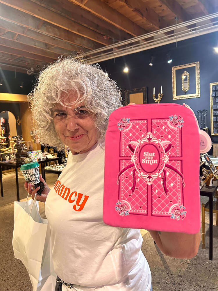 A woman with curly grey hair wearing a white "romancy" t-shirt holds up a bright pink embroidered book pouch that reads "Slut FOR Smut" inside a boutique store.