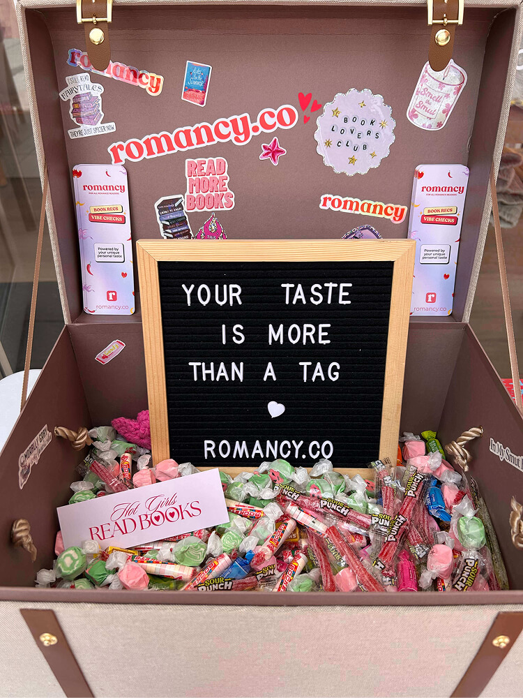 A open vintage-style suitcase used as a display filled with wrapped candies, sour punch straws, and a sticker reading "Hot Girls READ BOOKS". The open lid features stickers and a small black felt letter board that reads "YOUR TASTE IS MORE THAN A TAG - ROMANCY.CO".