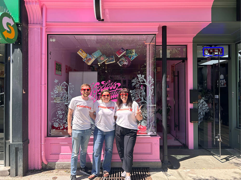hree people wearing white t-shirts with "romancy" printed in red, paired with matching red heart-shaped sunglasses, stand outside a bright pink bookstore storefront. Window decals read "Hopeless Romantic" alongside book illustrations.