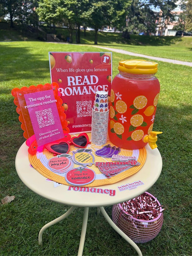 A small round white table set up outdoors on grass displaying a drink dispenser with red lemonade, stacks of paper cups, red heart sunglasses, promotional signs, and fruit-shaped stickers (peach, lemon, tomato, eggplant). A pink disco ball planter sits below the table.