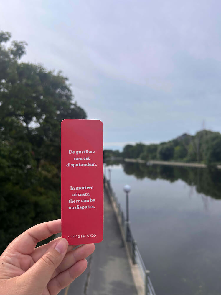 A hand holding a red bookmark outdoors with a river and trees in the background. The bookmark features the text "De gustibus non est disputandum. In matters of taste, there can be no disputes. romancy.co".