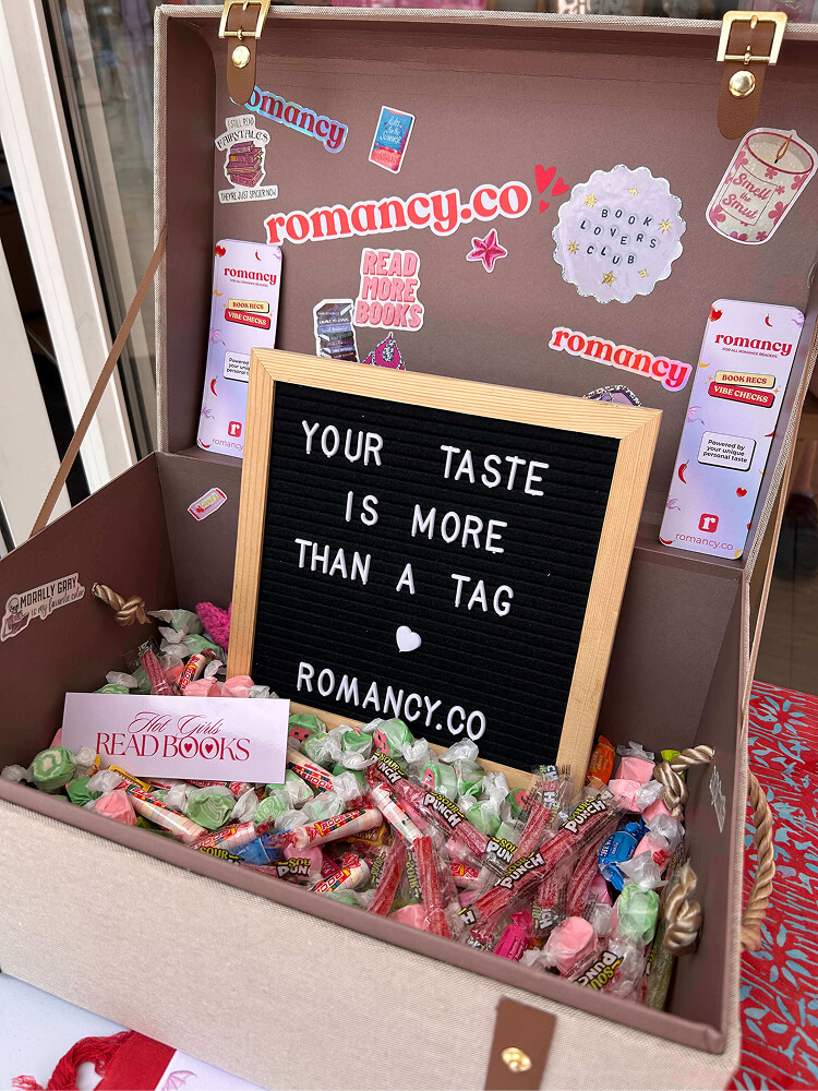 A open vintage-style suitcase used as a display filled with wrapped candies, sour punch straws, and a sticker reading "Hot Girls READ BOOKS". The open lid features stickers and a small black felt letter board that reads "YOUR TASTE IS MORE THAN A TAG - ROMANCY.CO".